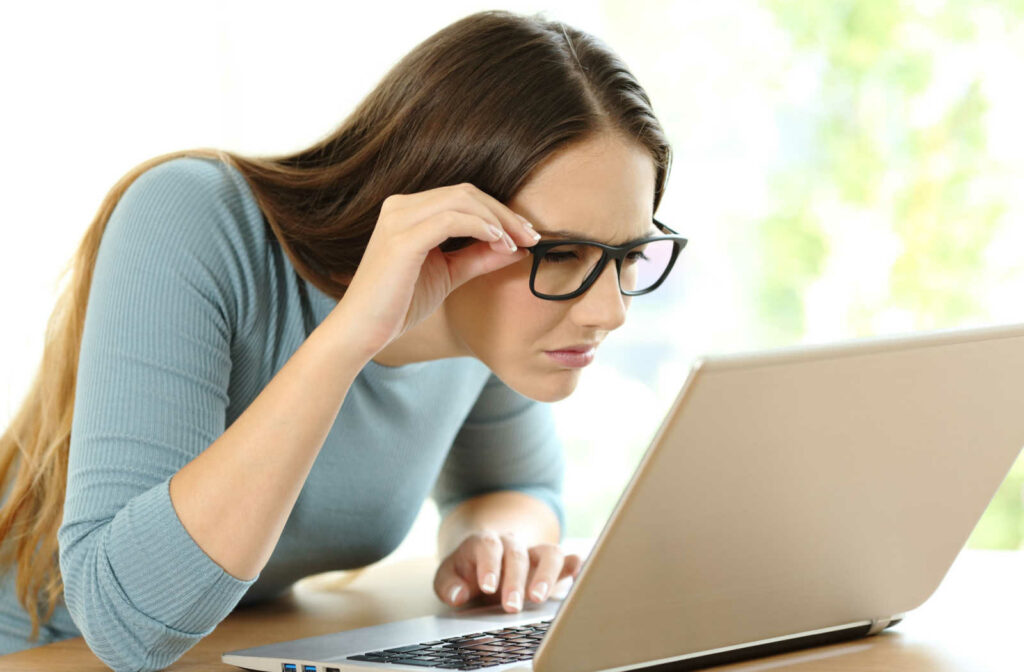A young female in a blue-colored long sleeve shirt and wearing black-colored frame eyeglasses is sitting in front of her computer and her face is too close to the computer screen.