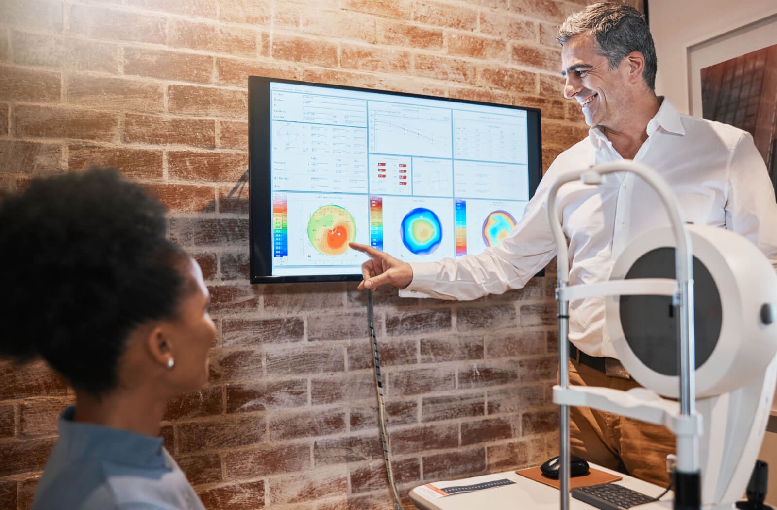 An excited optometrist pointing to a screen and presenting the findings of the eye exam to his patient.